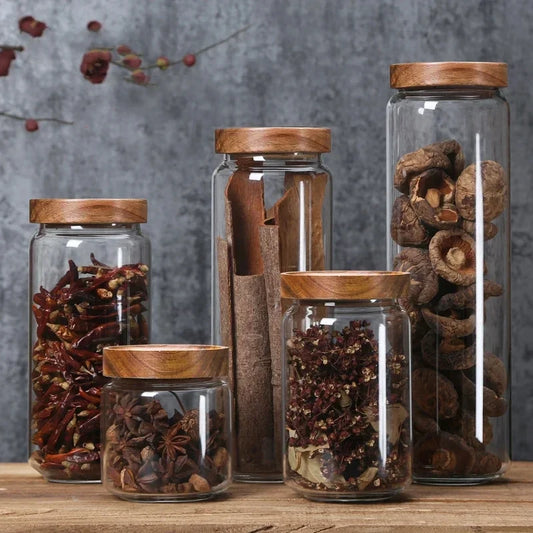 Six wooden-lid glass storage jars in a row, filled with dried chillies, cinnamon and mushrooms
